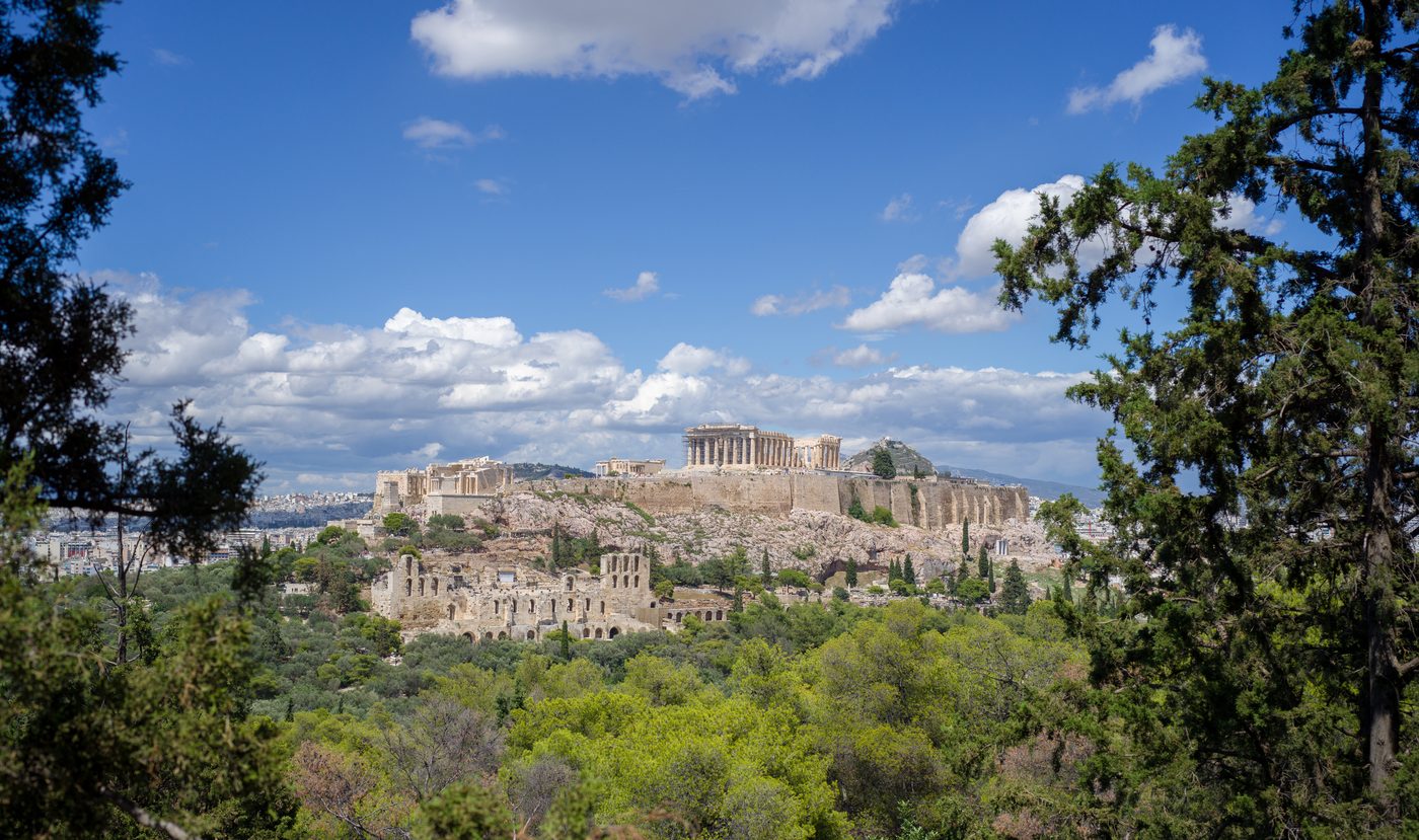Panoramic view of the Acropolis and Parthenon from Filopappou Hill in Athens, with green trees in the foreground and blue sky above