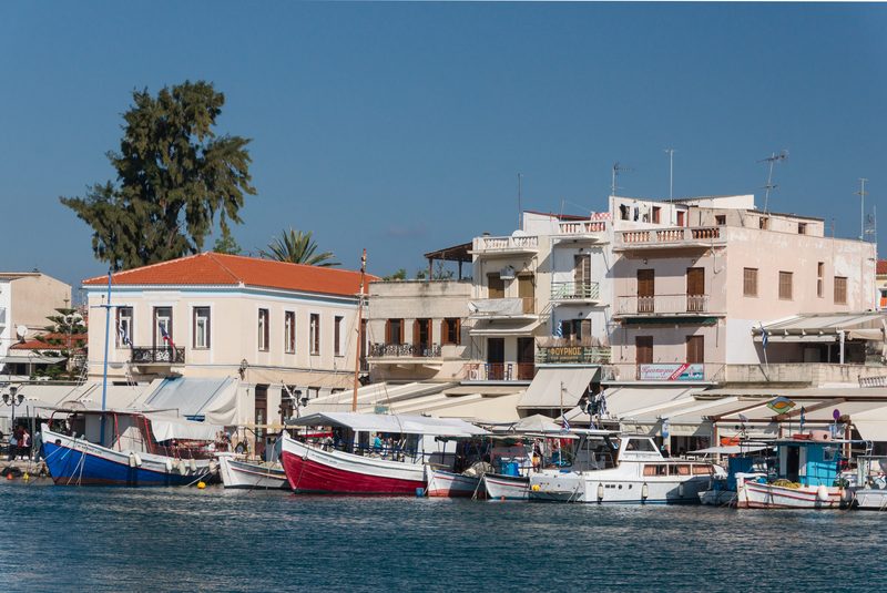 Fishing boats moored along the harbour embankment in Aegina town, Greece, with colourful waterfront buildings in the background