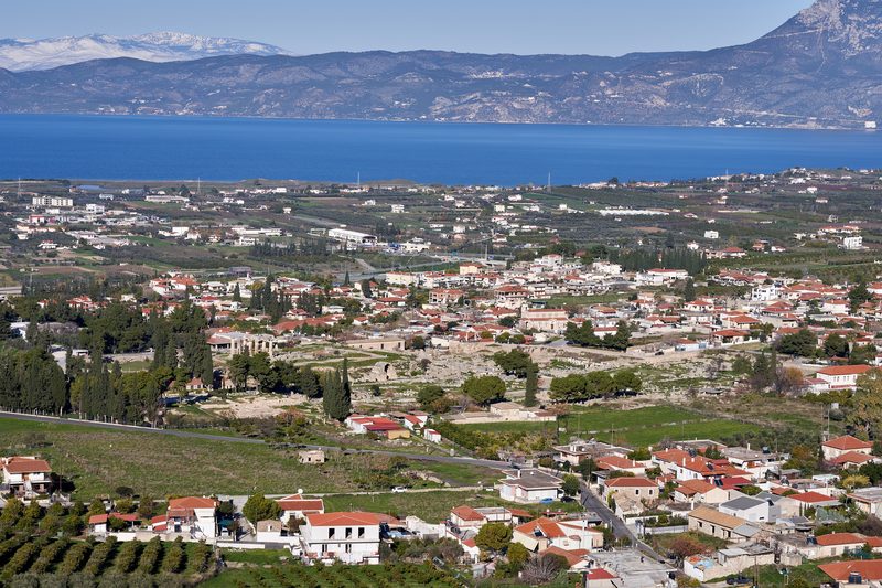 The archaeological site of Ancient Corinth seen from above, with scattered ruins of the Roman forum and the hill of Acrocorinth rising behind