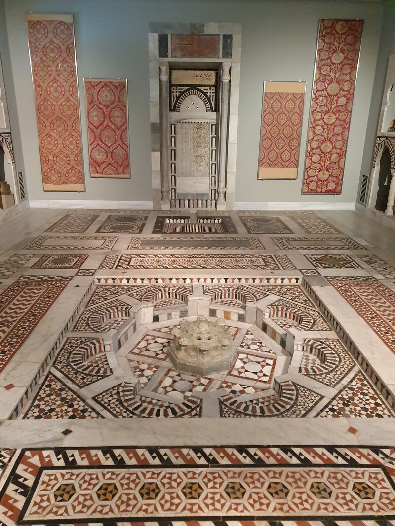An elaborately carved marble sarcophagus and inlaid stone decoration in a gallery at the National Archaeological Museum, with historic embroidered textiles displayed on the wall