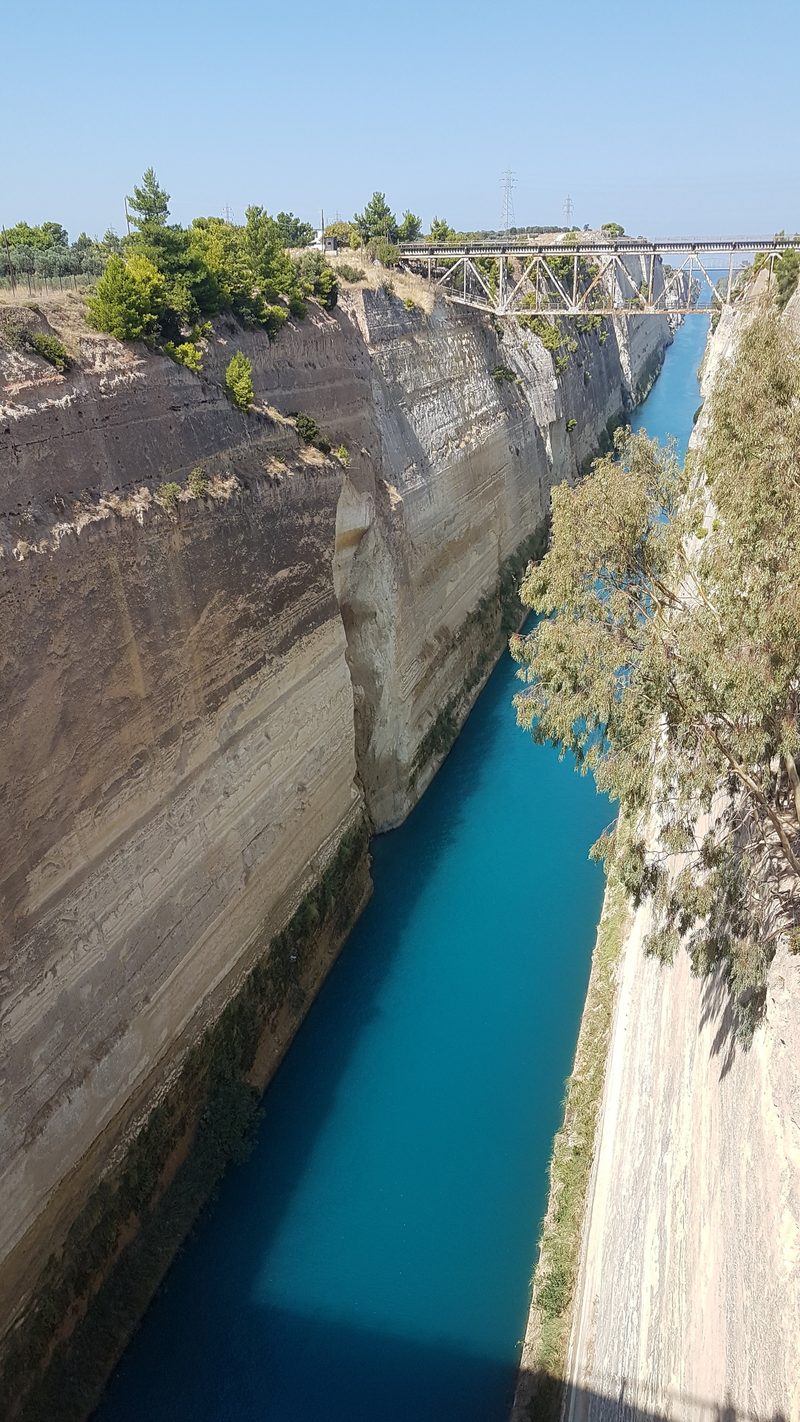 The steep sandy walls and turquoise water of the Corinth Canal, with the old railway bridge in the background