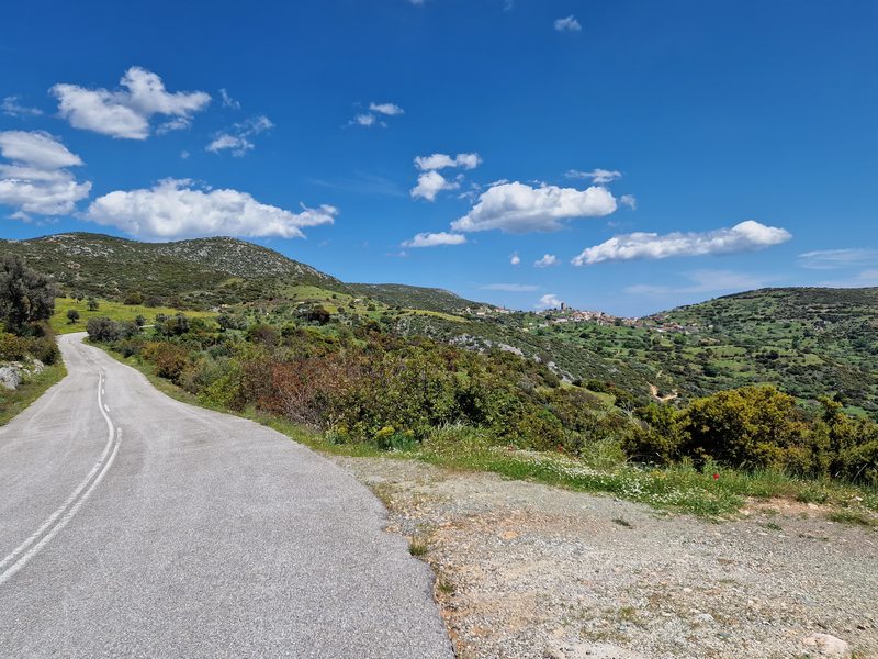 A quiet country road winding through the hills of Evia, with a small hillside village visible in the distance