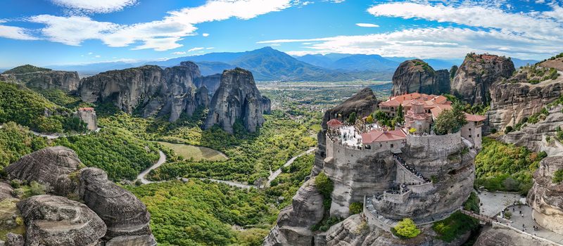 Panoramic view of three Meteora monasteries -- Rousanou, Varlaam, and Great Meteoron -- sitting atop towering sandstone rock pillars against a blue sky