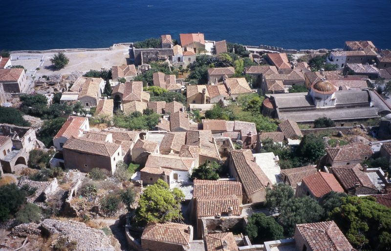Aerial view of Monemvasia's Lower Town showing medieval stone buildings and churches clustered behind fortress walls with the sea beyond
