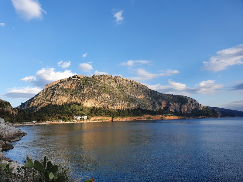 The rocky crag of Palamidi fortress rising above a small beach on the Nafplio coastline, seen across calm water