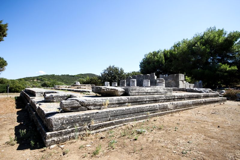 Stone foundations of the ancient Temple of Nemesis at Rhamnous archaeological site, showing the Doric temple base amid dry grass and scattered ruins