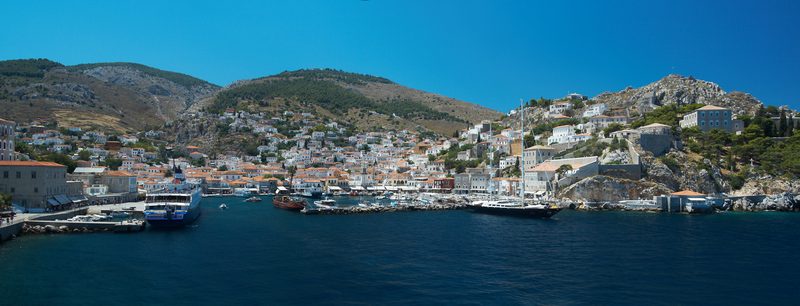 Panoramic view of Hydra harbour in Greece showing the crescent-shaped port lined with stone mansions rising up the hillside, boats moored in calm blue water