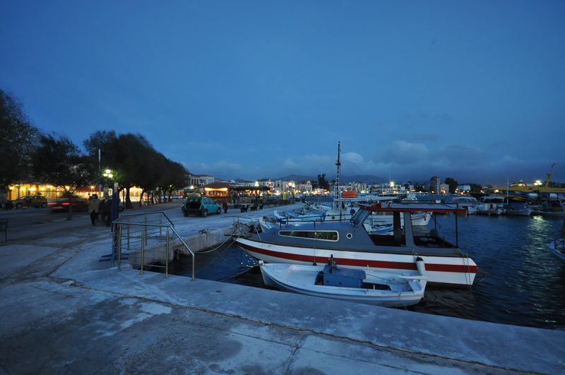 Aegina harbour at dusk with colourful fishing boats in the foreground, waterfront tavernas and neoclassical buildings along the quay, and a warm evening sky
