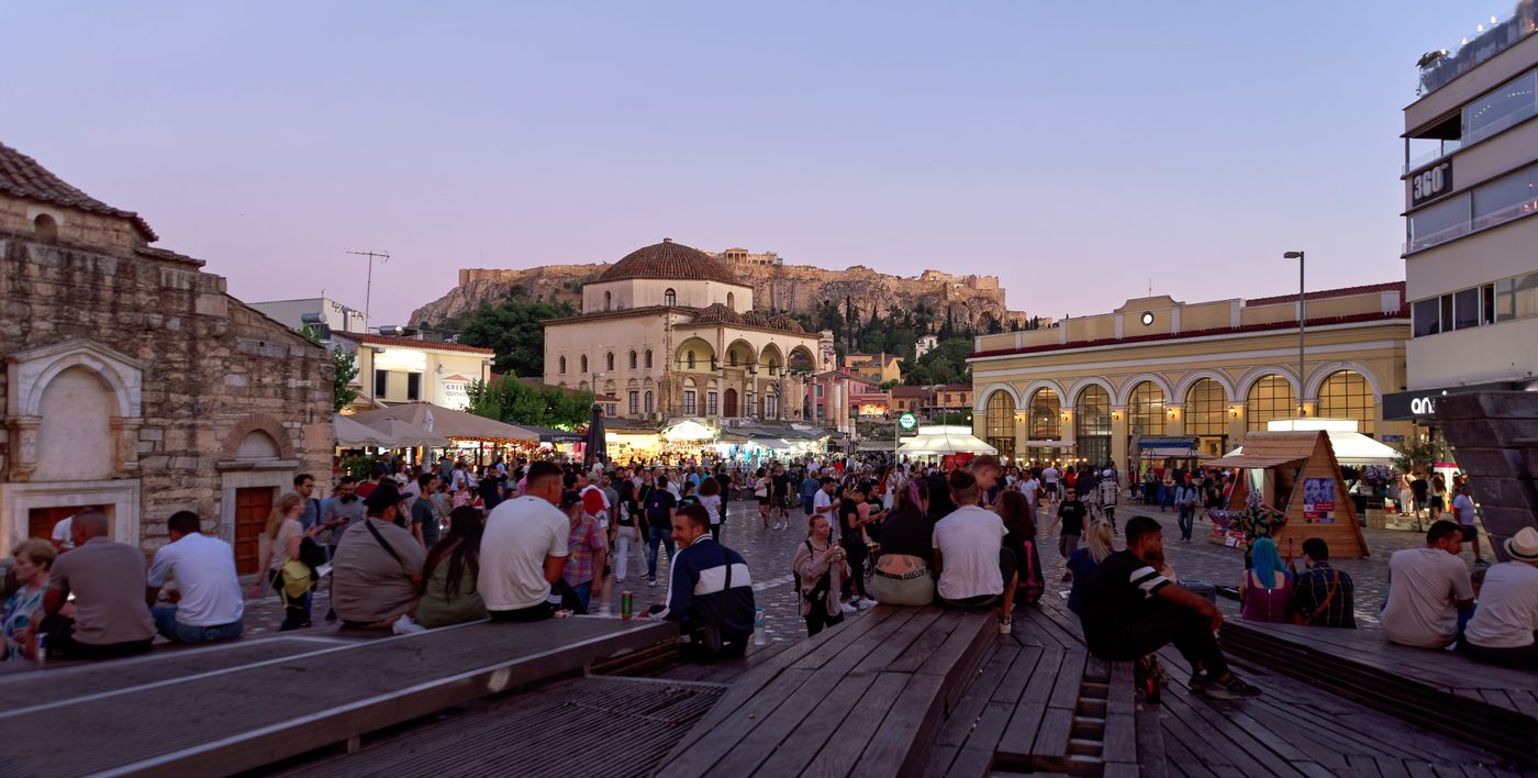 Monastiraki Square in Athens bustling with visitors on a warm summer evening, with illuminated historic buildings and the Acropolis visible in the background