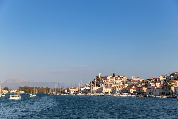 Poros town waterfront seen from the sea with white and terracotta-roofed buildings rising to the clock tower, yachts in the harbour and mountains beyond
